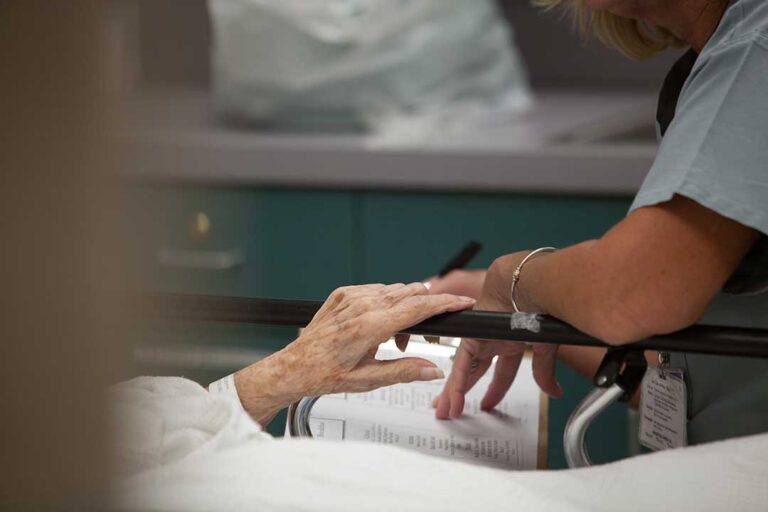 Older adult using a walker along a bright assisted living hallway while staff member provides support.